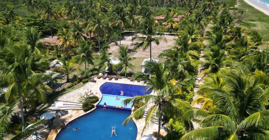 All inclusive em Natal: como aproveitar o resort sem deixar de conhecer o destino Área com piscina rodeada por palmeiras e pessoas relaxando ao sol.