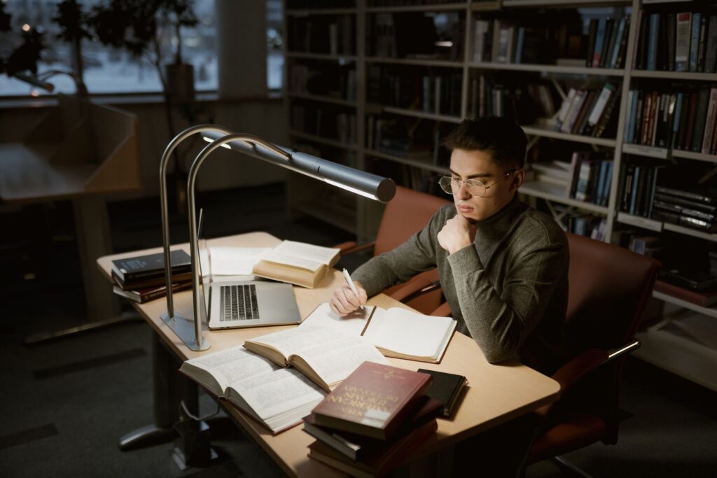 Como estudar para concurso trabalhando o dia inteiro Candidato estudando para concurso em mesa de biblioteca, com livros abertos, caderno e notebook sob a luz de uma luminária